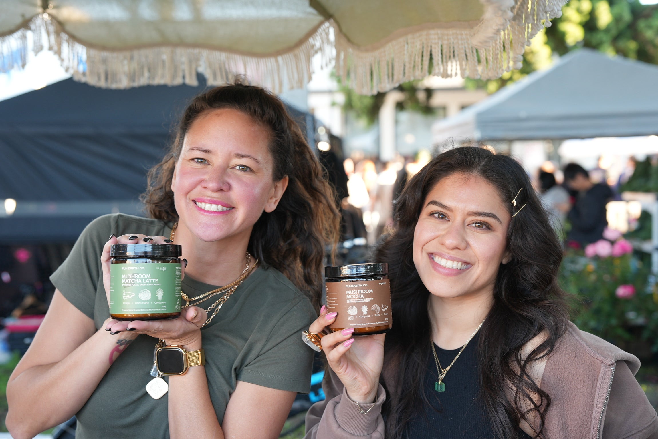 Two women holding Fun Earth Mushroom Matcha Latte and Mushroom Mocha jars at a community market